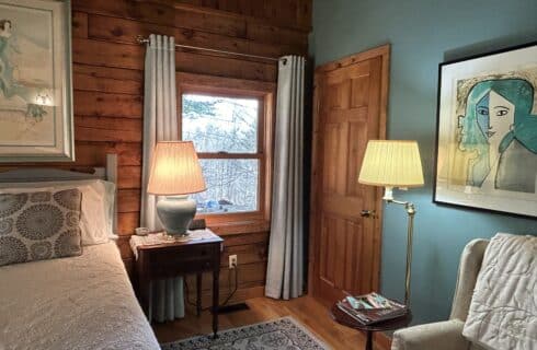 The bed has a white quilted spread with aqua patterned pillows. The nightstand has a turquoise lamp on a doily. The wingback chair is off-white with a velvet throw. The window looks out over the fire pit.