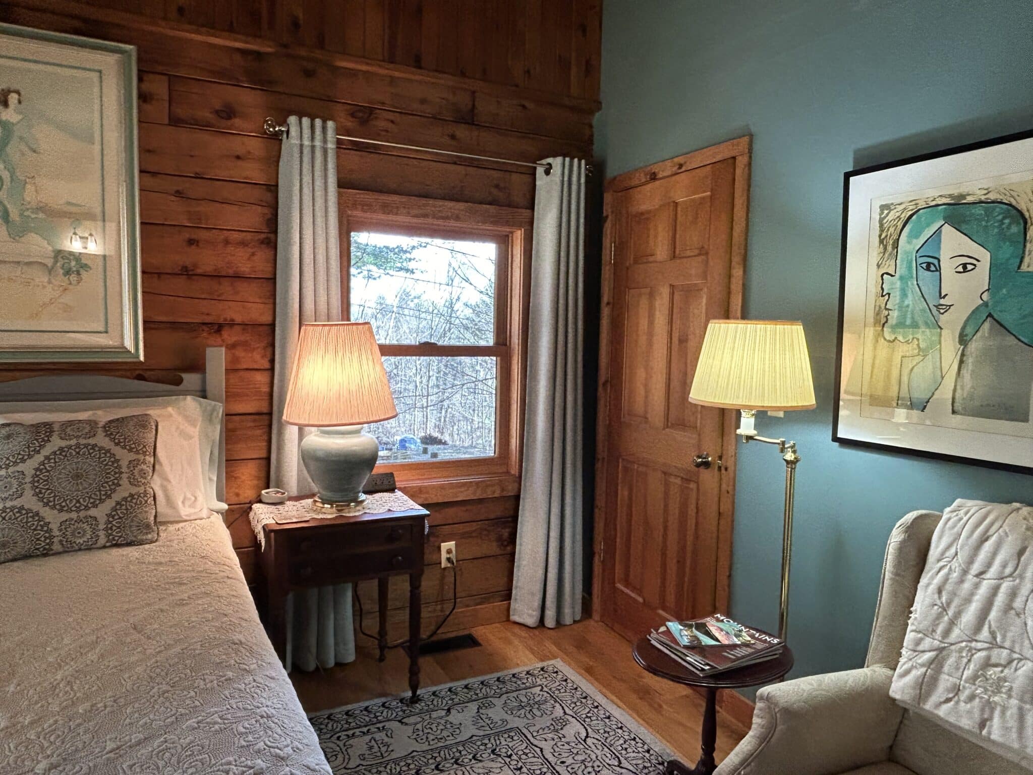 The bed has a white quilted spread with aqua patterned pillows. The nightstand has a turquoise lamp on a doily. The wingback chair is off-white with a velvet throw.  The window looks out over the fire pit.