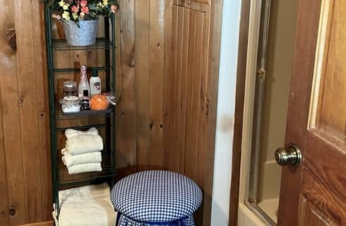 The Shared bath has a shelving unit with extra towels and amenities. The is a stool covered in gingham fabric.