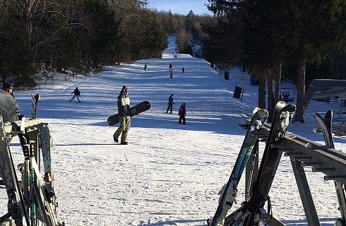 Narrow ski hill flanked by trees with skiers and snowboarders on a sunny day