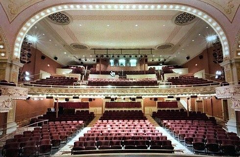 Inside of a small theatre with red seats, balcony and curved decorative ceiling