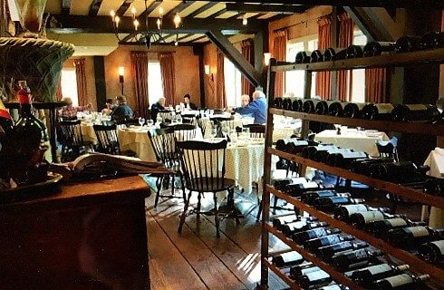 Rustic restaurant with wood beam ceiling and black chandeliers with dining tables next to a wine rack full of different bottles of wine