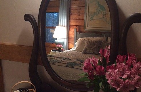 Large oval mirror flowers beside reflecting a king size bed with large painting overhead and side table with lamp near a window