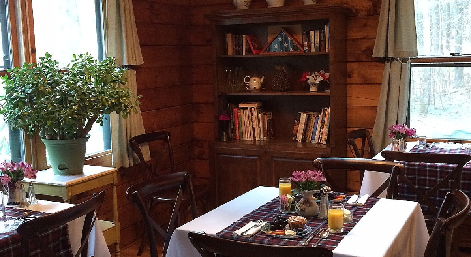 Corner of a room with wood paneled walls with three dining tables set for breakfast, bookshelf and table with plant in front of a bright window