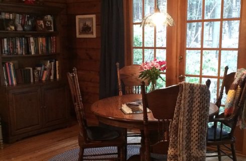 Wood dining table with four chairs under small hanging light next to French doors and tall bookshelf filled with books