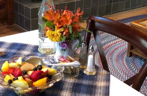 White table with blue plaid cloth holding dish of fruit salad, vase of fresh flowers, salt and pepper and dish of sugar packets