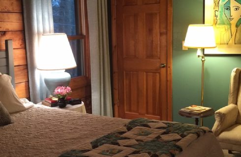 King bed with white quilt, side table with lamp, books and flowers and reading chair in a room with green walls and one window and door