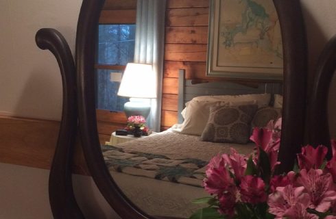 Oval antique mirror reflecting a king bed with blue headboard, white quilt and blue accent pillows wth side table and lamp