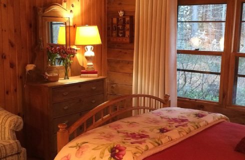 Bed with curved wooden footboard next to an upholstered chair, dresser with mirror and large window with white curtains