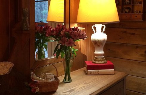 Wood dresser with white lamp on two stacked books, vase of pink flowers and basket of gift items with mirror behind