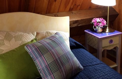 Double sized bed with a blue quilt and accent pillows and small side table holding a lamp and cup of flowers