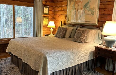 The Greylock Room with king size bed, nightstands with lamps, big window view of forest, large artwork above the bed, and area rug over hardwood oak floor.