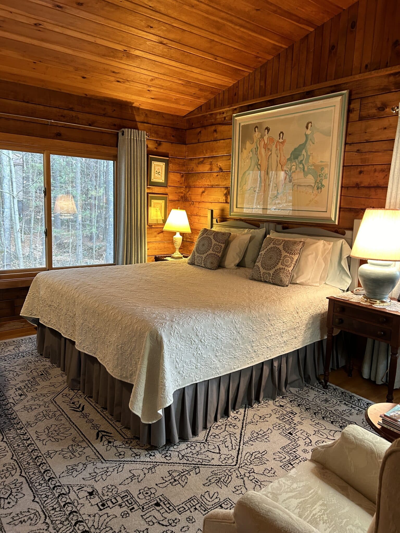The Greylock Room with king size bed, nightstands with lamps, big window view of forest, large artwork above the bed, and area rug over hardwood oak floor.