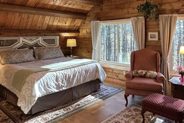 The Ramblewood Room has a king size bed, repurposed headboard, off-white spread, and a sitting area with 2 wingback chairs. The large windows with beige drapers look out over the front forest.