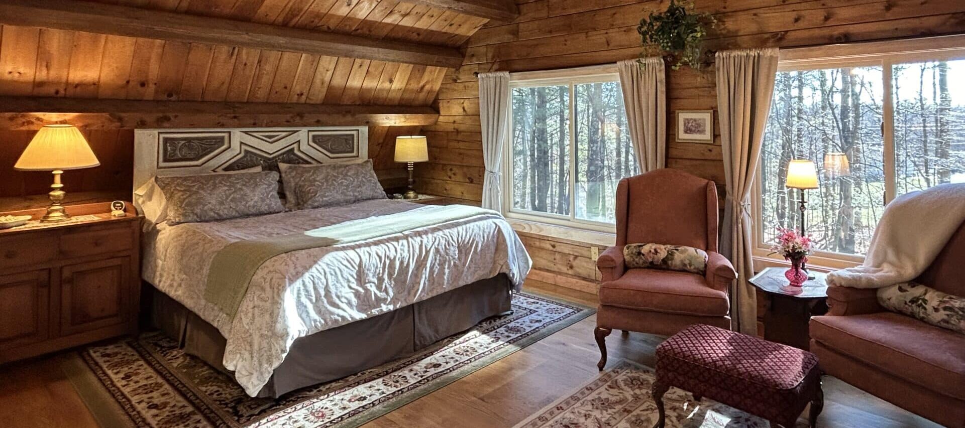 The Ramblewood Room has a king size bed, repurposed headboard, off-white spread, and a sitting area with 2 wingback chairs. The large windows with beige drapers look out over the front forest.