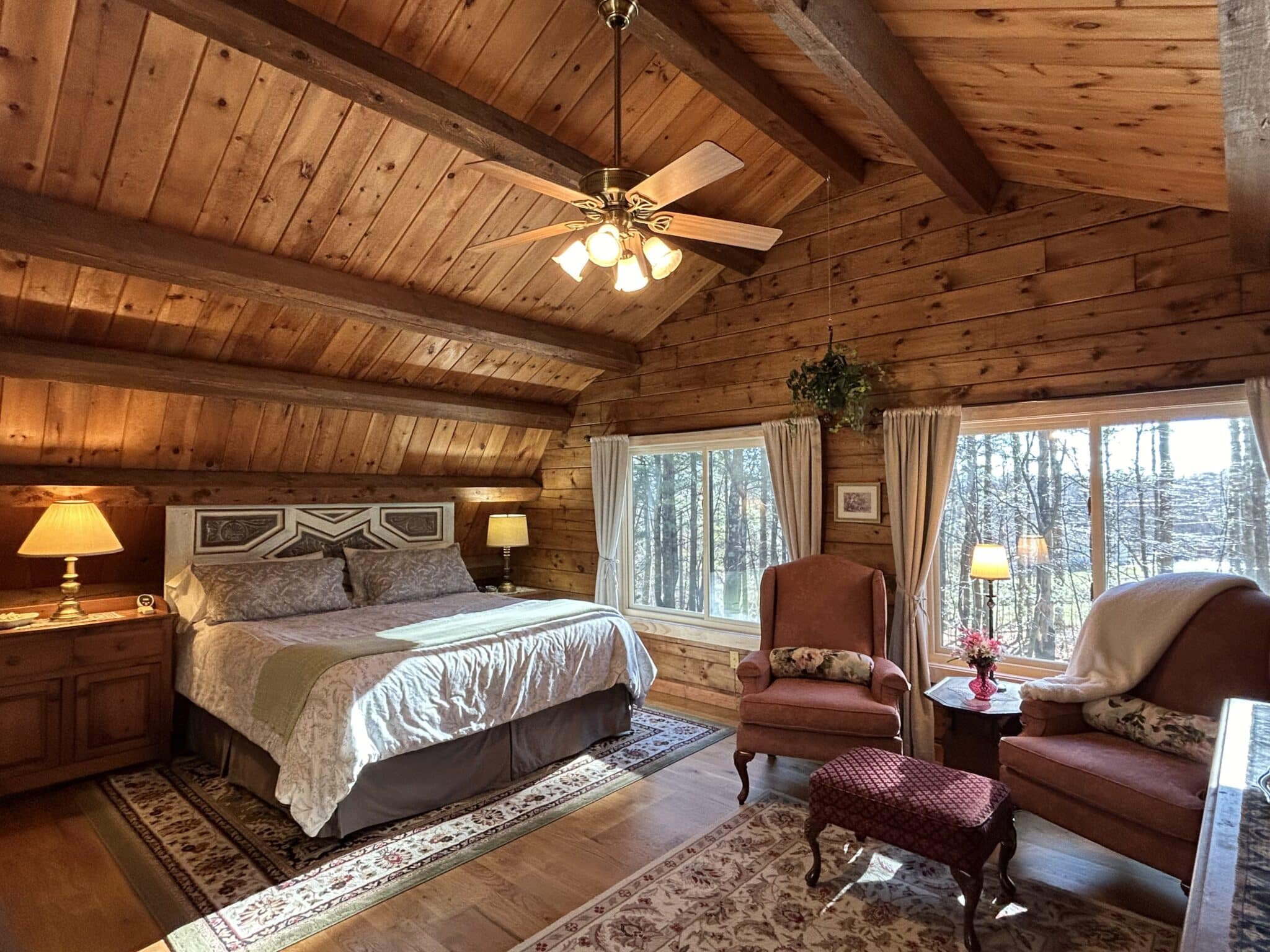 The Ramblewood Room has a king size bed, repurposed headboard, off-white spread, and a sitting area with 2 wingback chairs.  The large windows with beige drapers look out over the front forest.