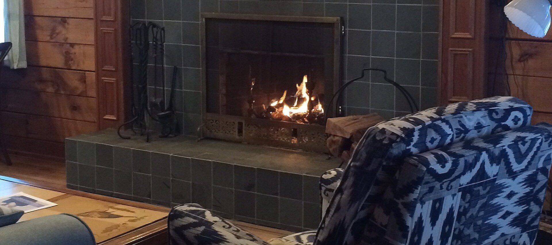 Fire roaring in a grey tiled fireplace with table, couch and sitting chair in a wood paneled room