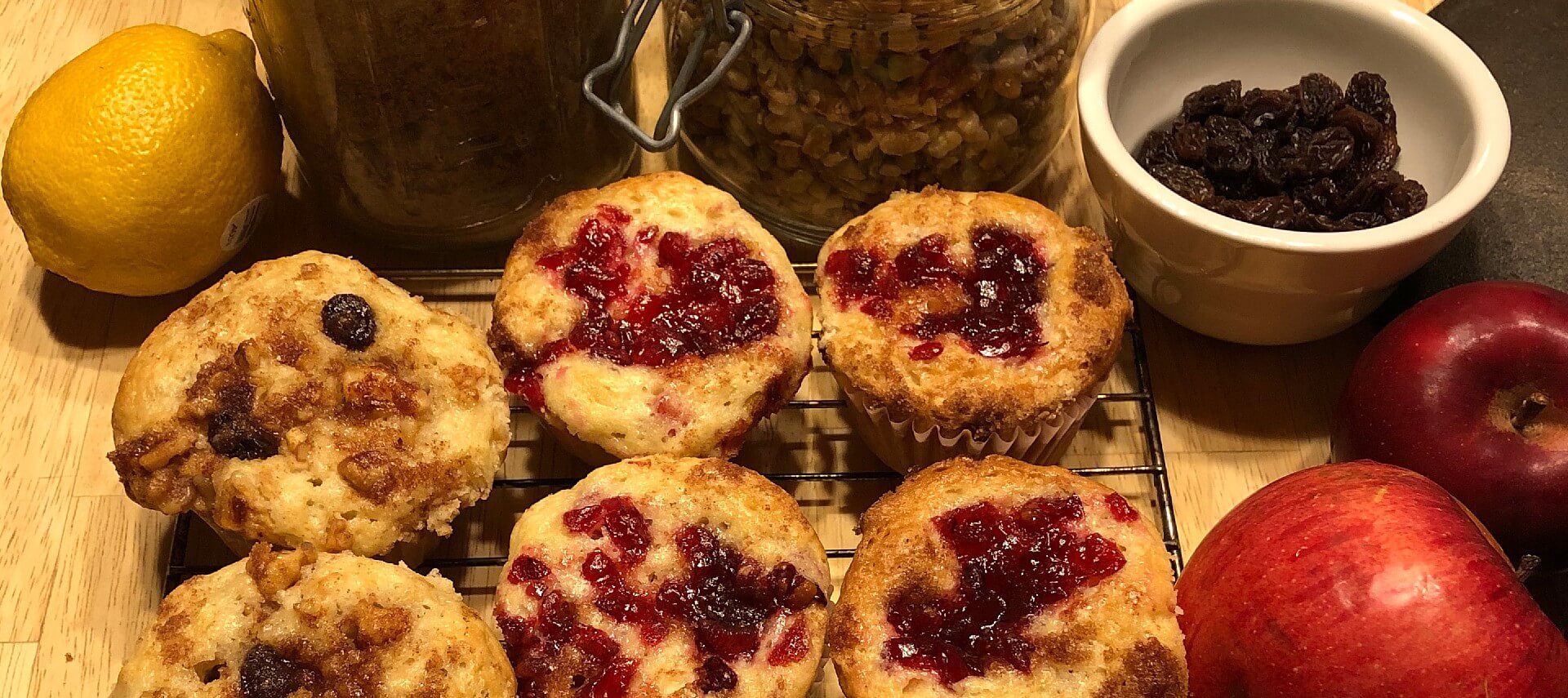 Six freshly baked muffins on a wire rack next to apples, bowl of raisins and jar of nuts