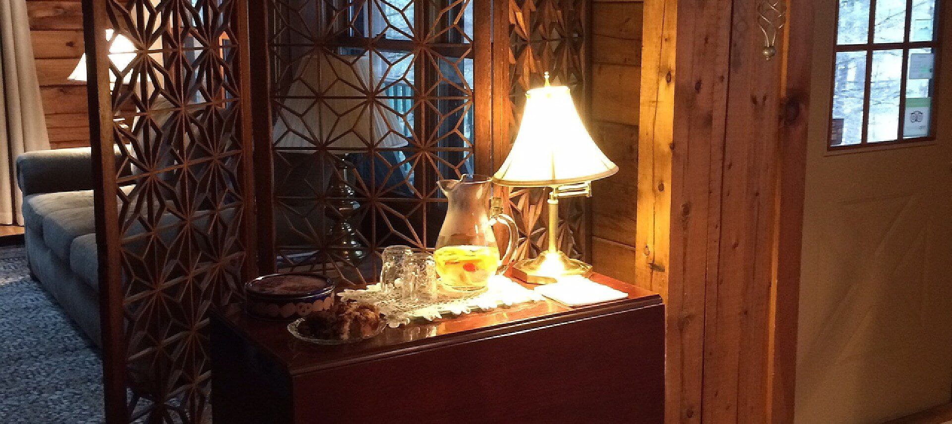 Wood table by a front door holding plate of cookies, pitcher of lemonade, glasses and lamp in front of a tall decorative screen