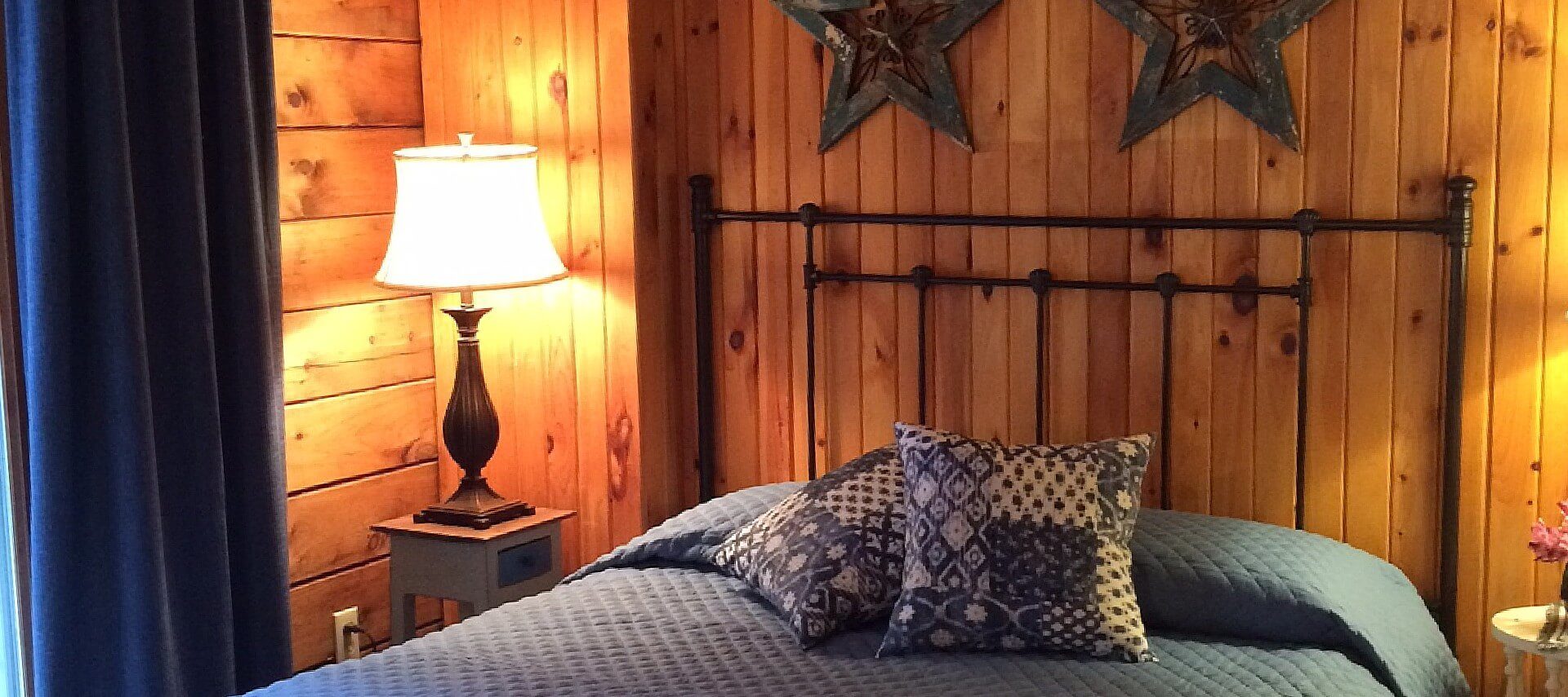 Queen bed with black wrought iron headboard and blue quilt, two side tables with lamps in a room with wood paneled walls