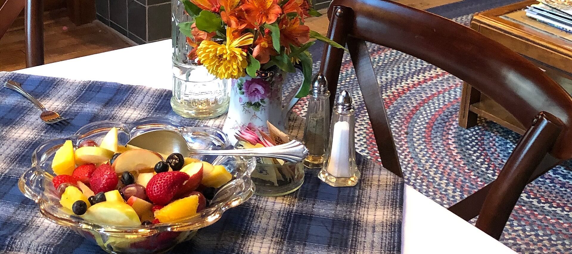 Table with blue plaid cloth holding glass bowl of fruit, vase with flowers, salt and pepper and dish of sugar packets