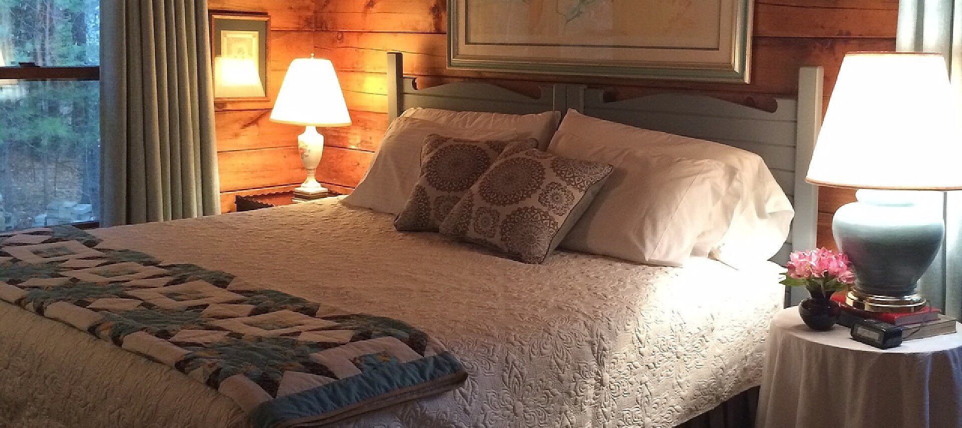 King bed with beautiful white quilt and coverlet, two side tables with lamps next to a large window with curtains