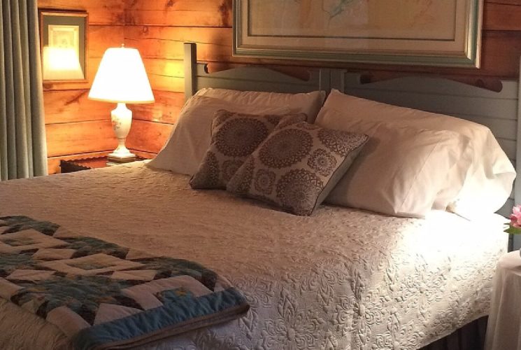 King bed with beautiful white quilt and coverlet, two side tables with lamps next to a large window with curtains