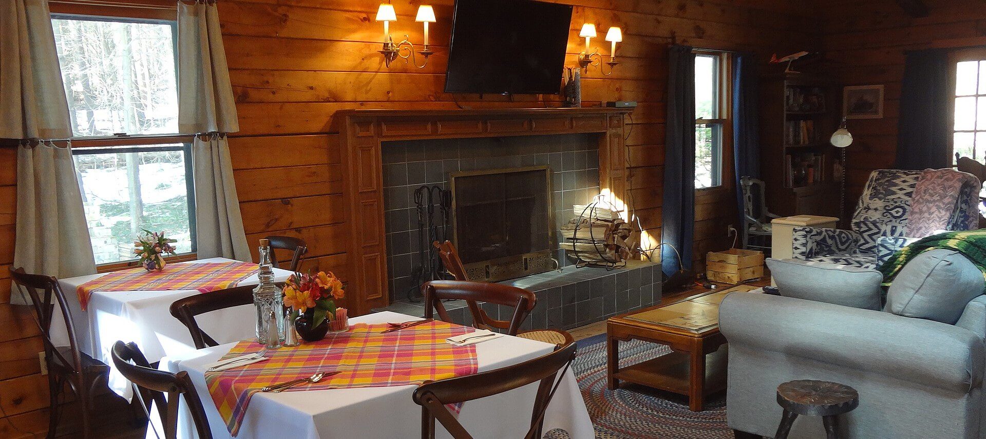 Spacious room with wood paneled walls, couch and chair in front of a fireplace with TV overhead and two dining tables