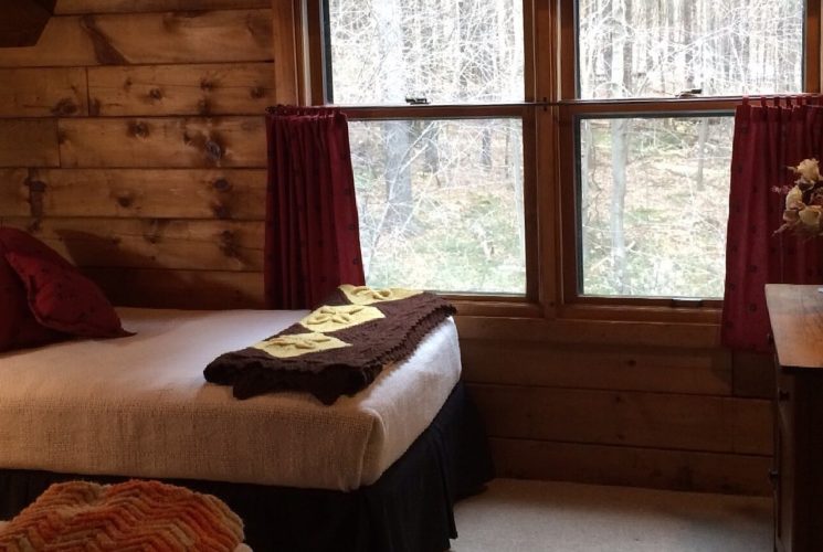 Twin bed with white quilt and red pillows underneath a large window with red curtains and views into a wooded area