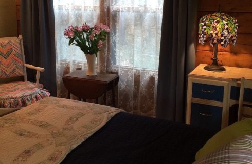 Bed with white flowered quilt, chair in the corner, window with lace curtains and white and blue reading desk holding a small tiffany lamp