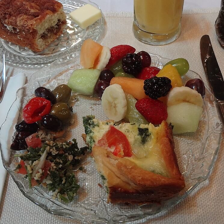 Round glass dish with olives, breakfast quiche, fruit salad next to glass of orange juice and plate of coffee cake