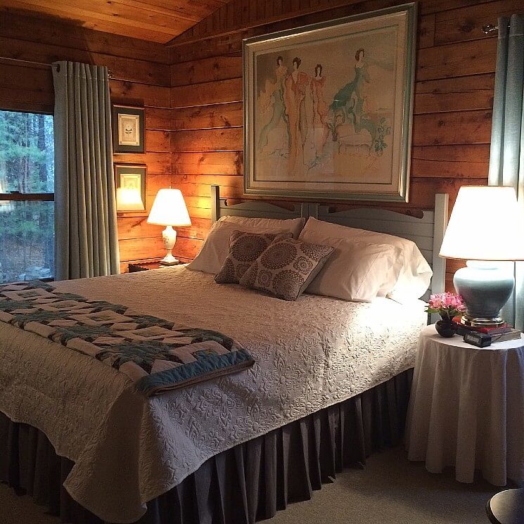 King bed with beautiful white and blue quilts, side tables with lamps and large painting above in wood paneled room