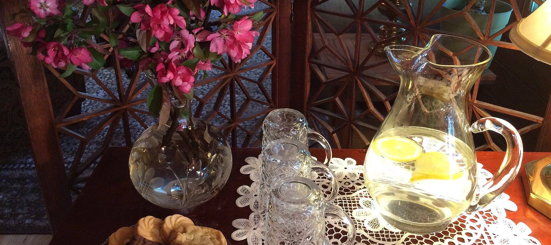 Dark wood table with lace doily placemat, three glass cups, pitcher of water with sliced lemons and vase with pink flowers