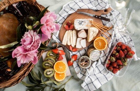 Board of cheese and fruit on a white and black checkered napkin net to a wooden basket with pink flowers