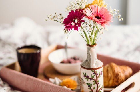 Wooden tray on a bed holding cup of coffee, croissant, bowl of fruit and vase with daisies