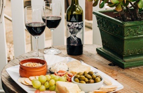 White plate of fruit, cheese and crackers next to bottle and two glasses of red wine on a brown table